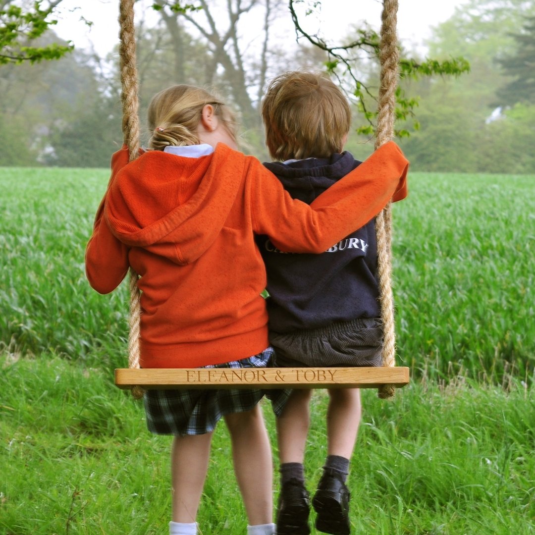 Two children on an oak and rope swing with 'Eleanor & Toby' engraved, standing in a grassy field.