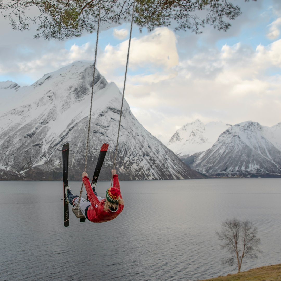 Person on an oak and rope swing with skis over a lake and snowy mountains