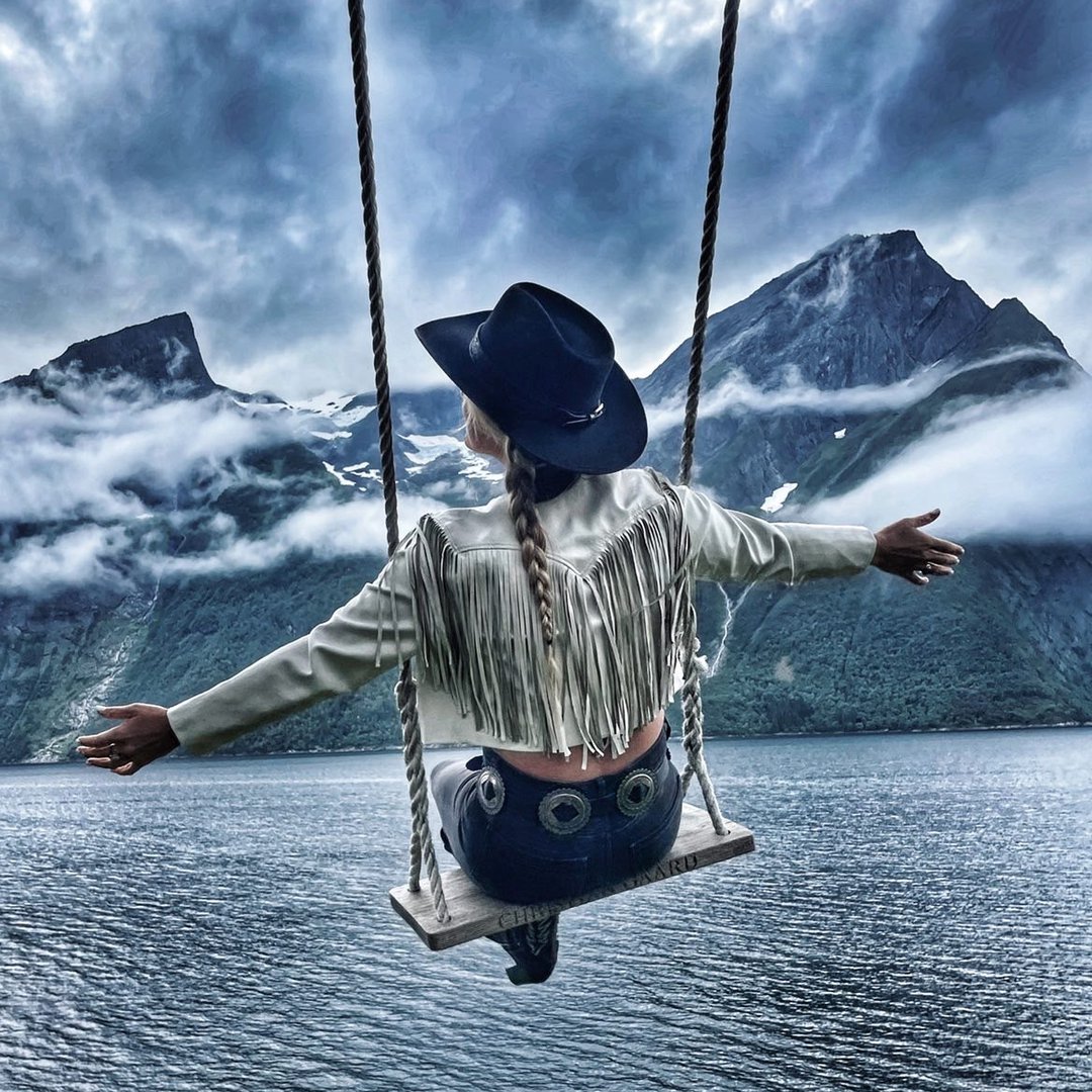 Person in a cowboy hat and fringed jacket on an oak and rope swing with mountains and lake in the background