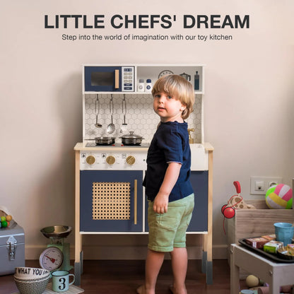 Child standing next to a toy kitchen set with various accessories on a wooden floor.
